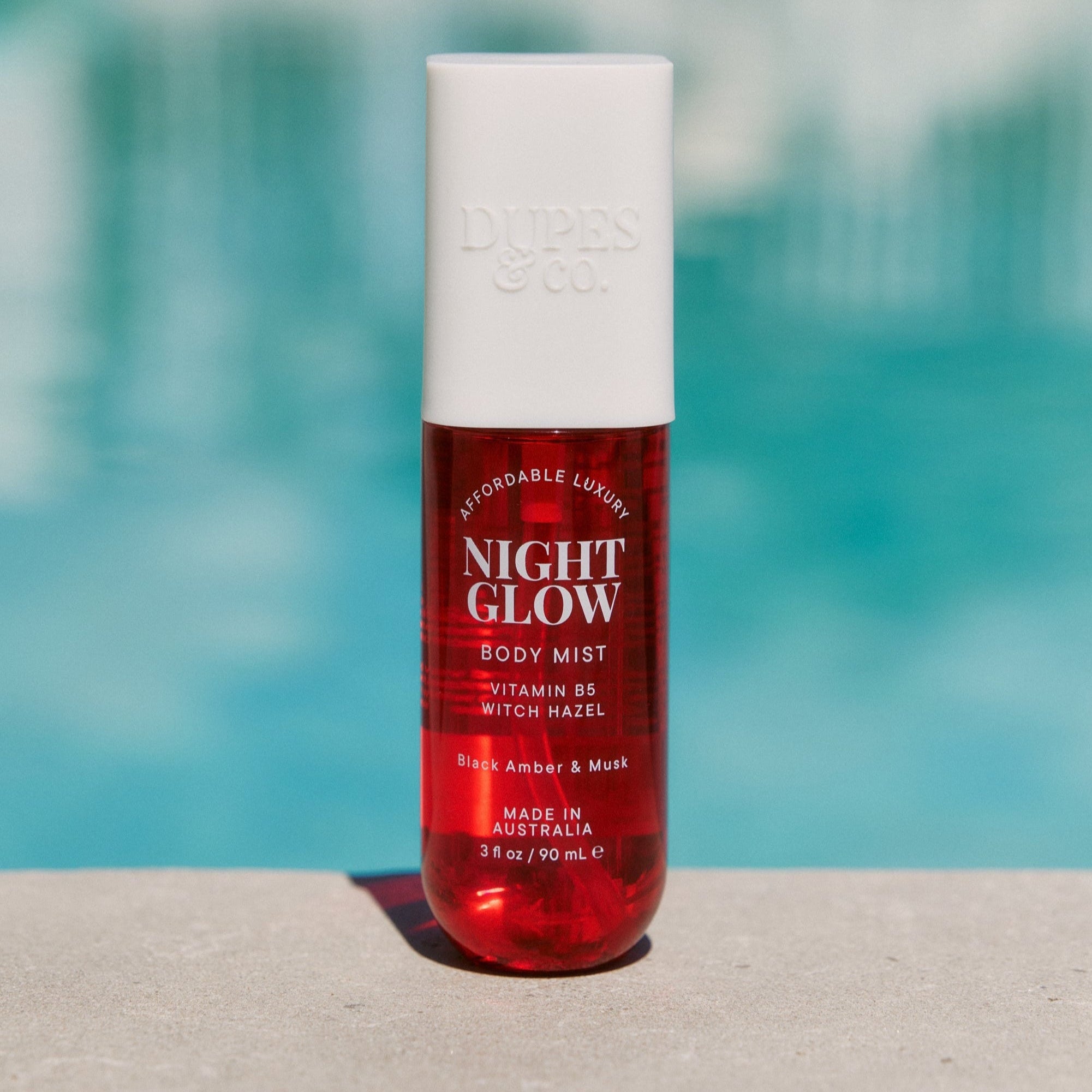A red bottle of 'Night Glow Black Amber & Plum Body Mist' with a white cap, displayed against a poolside background.