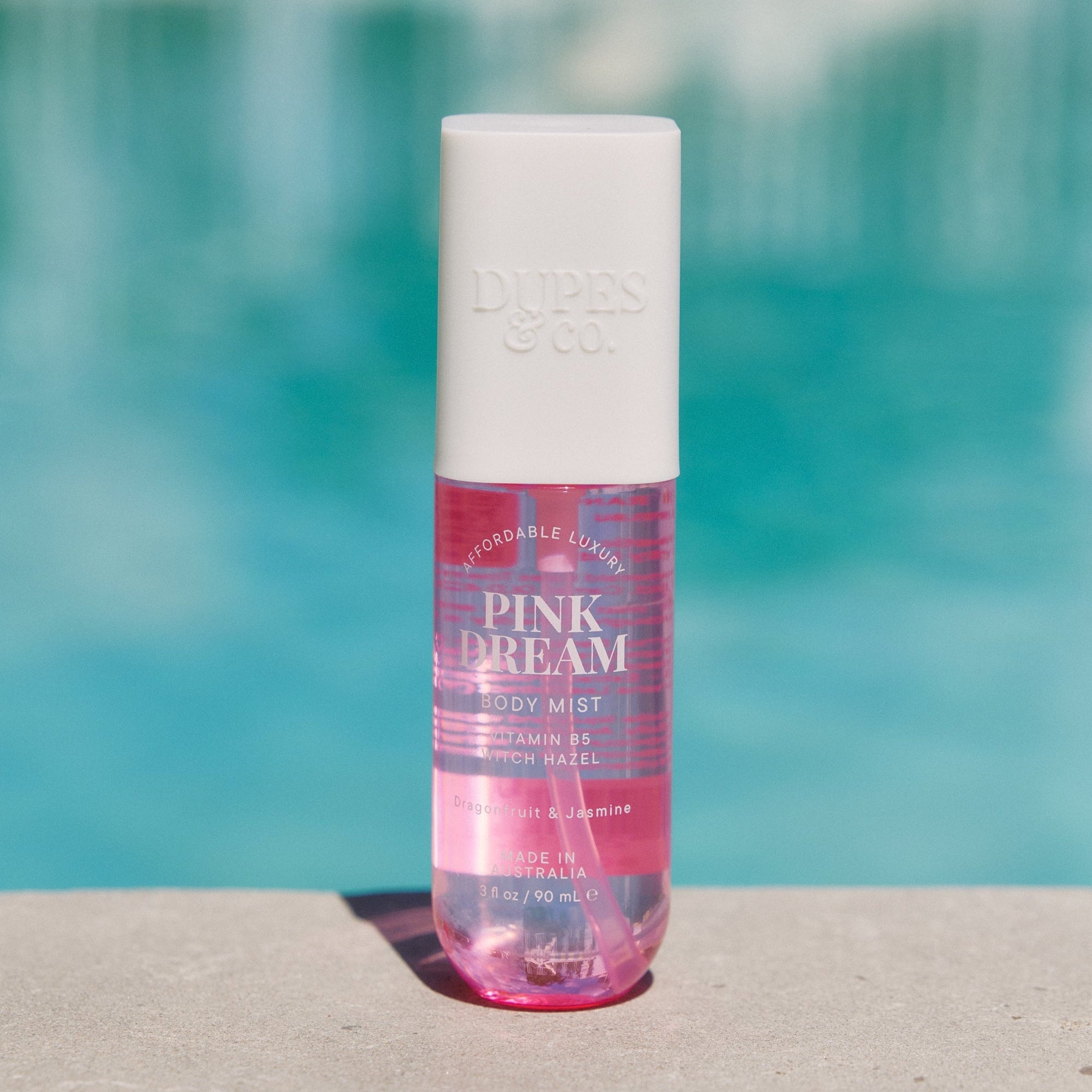 A bottle of Pink Dream body mist with pink liquid visible, placed on a surface with a pool in the background.
