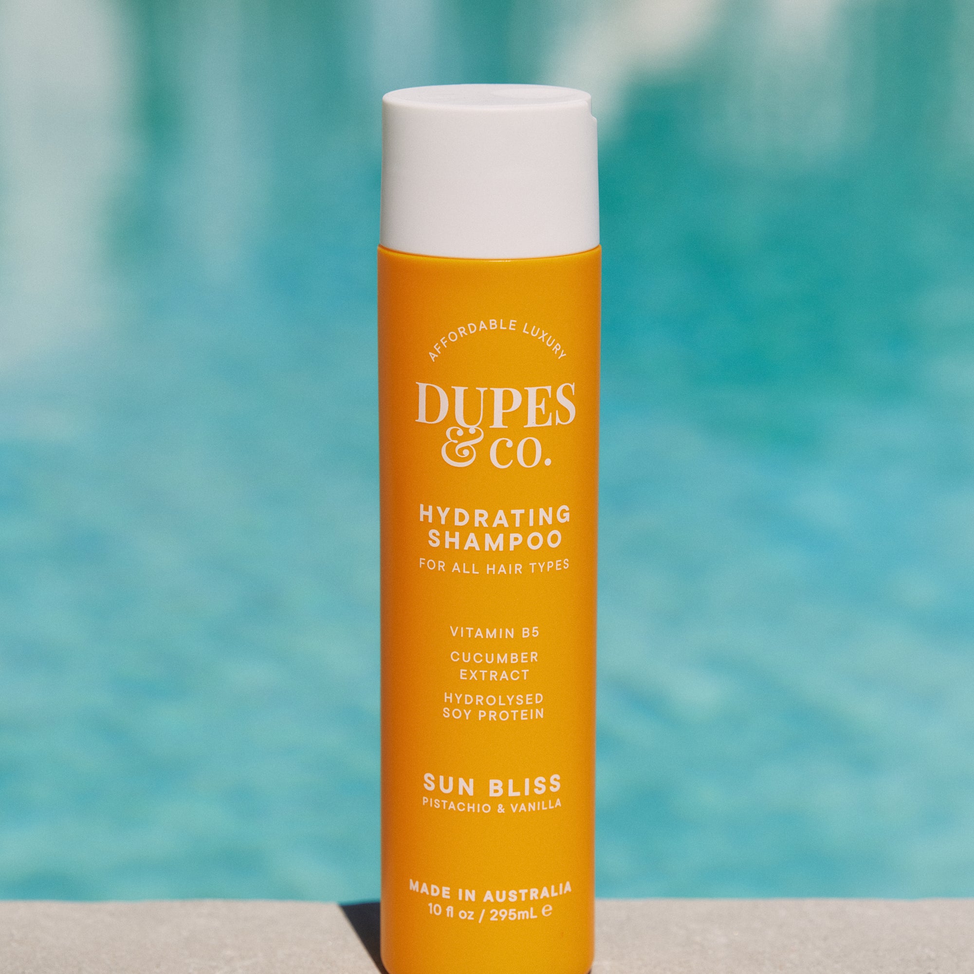 Orange shampoo bottle with white cap labeled 'Dupes & Co.' in front of a blurred pool background