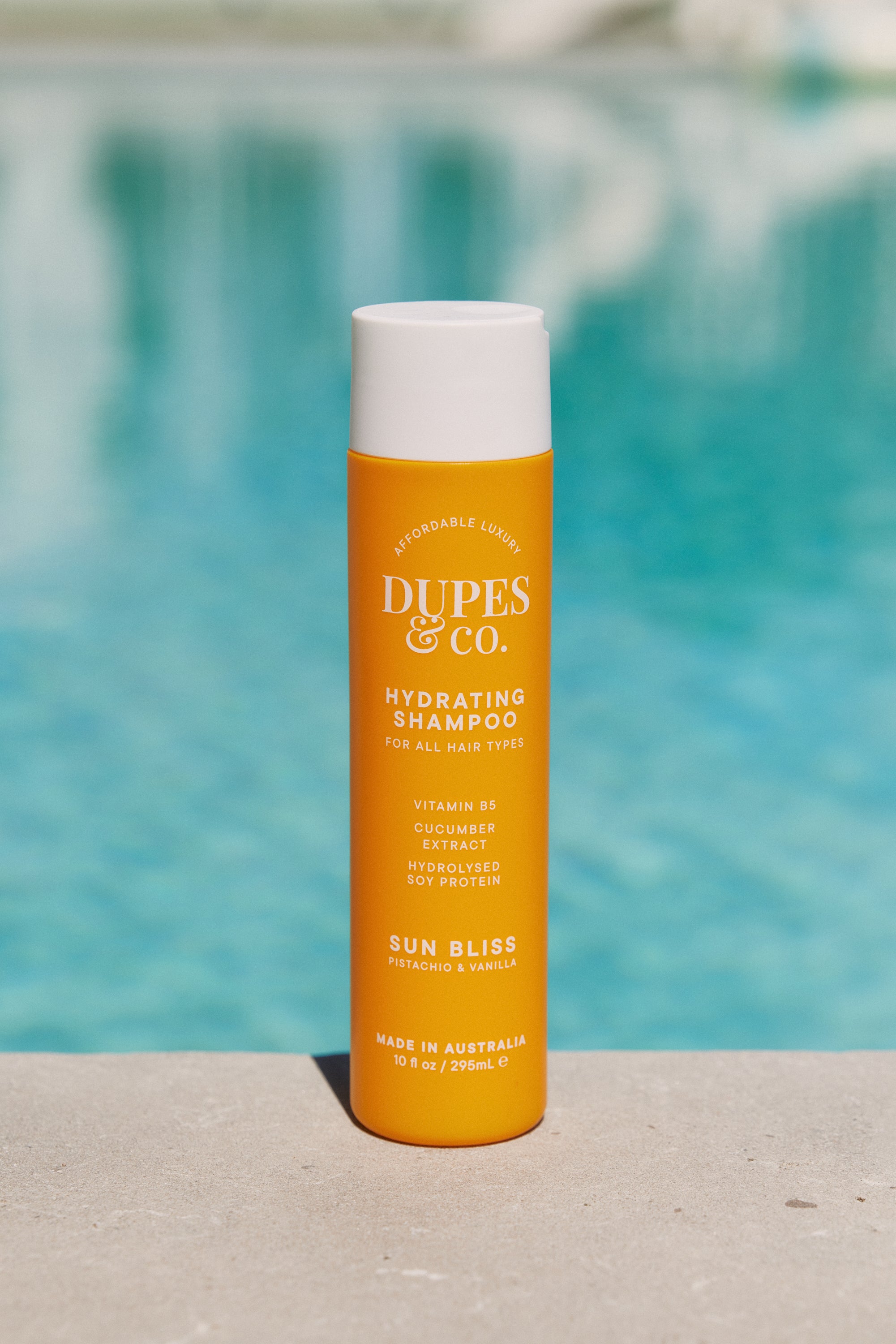 Orange shampoo bottle with white cap labeled 'Dupes & Co.' in front of a blurred pool background